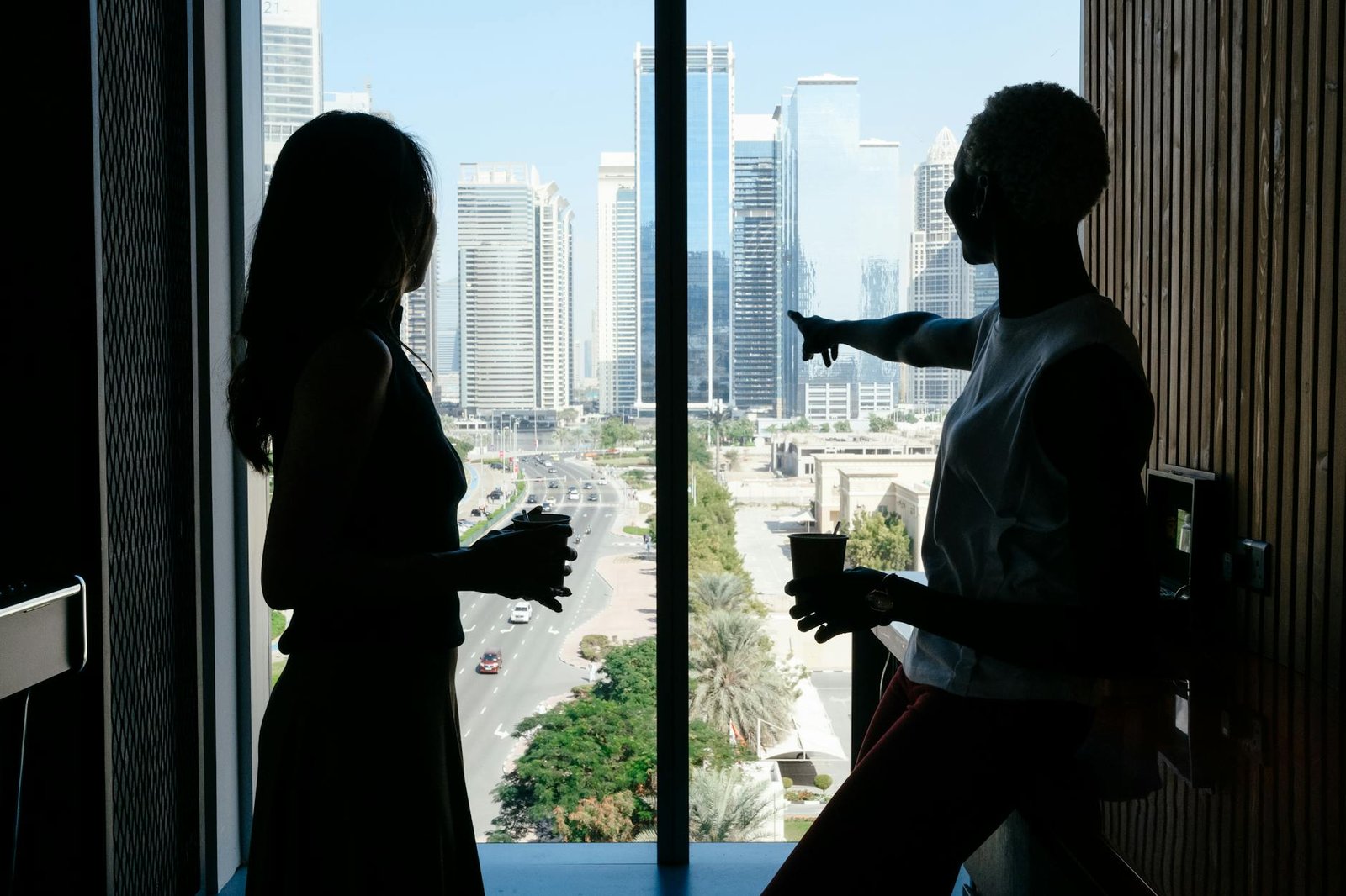 Side view of anonymous diverse business partners with coffee talking about job while admiring tall contemporary buildings of large megapolis