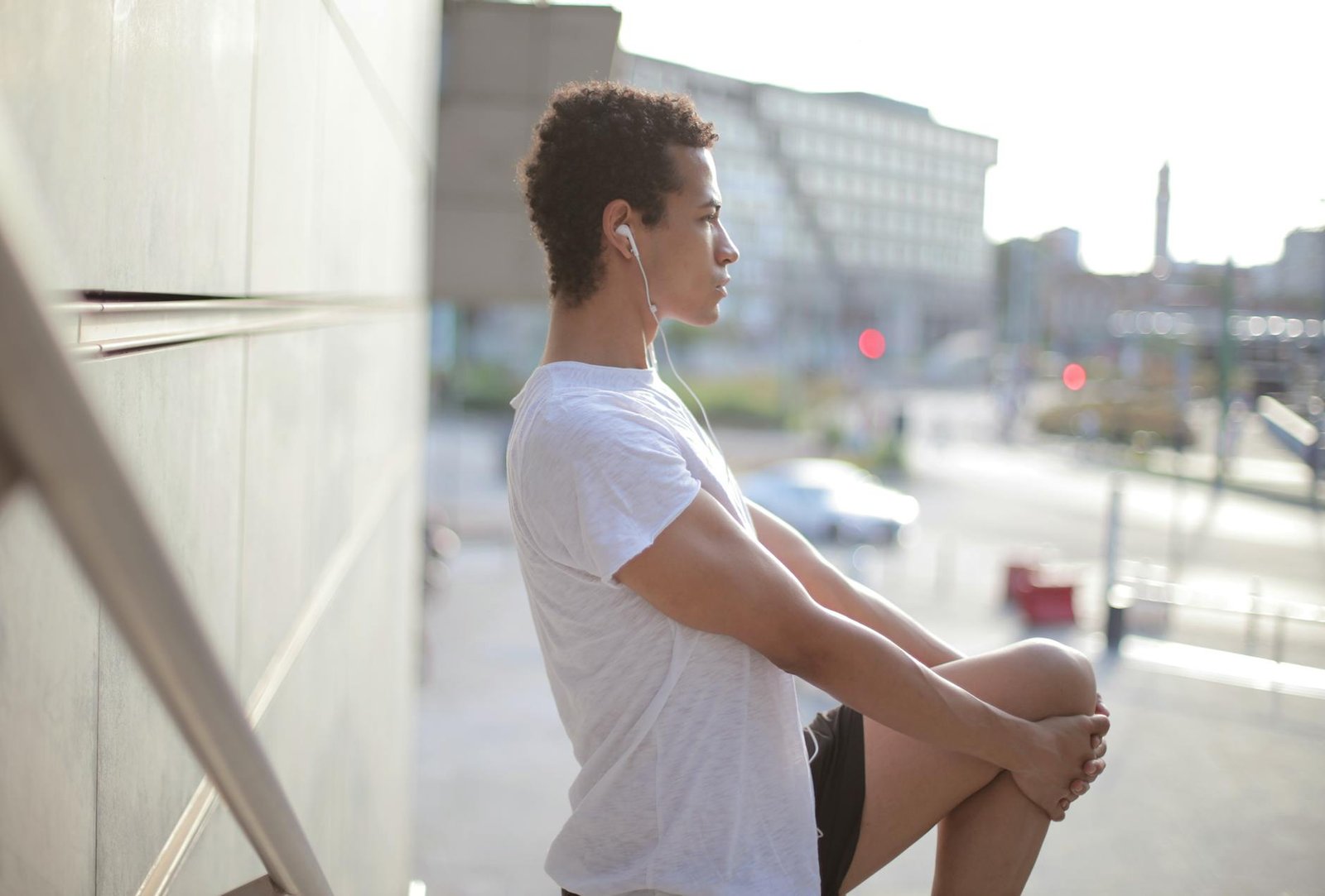 Side view of calm thoughtful African American male athlete in earphones and sportswear looking away and stretching leg while standing alone and listening to music on street near modern building in city